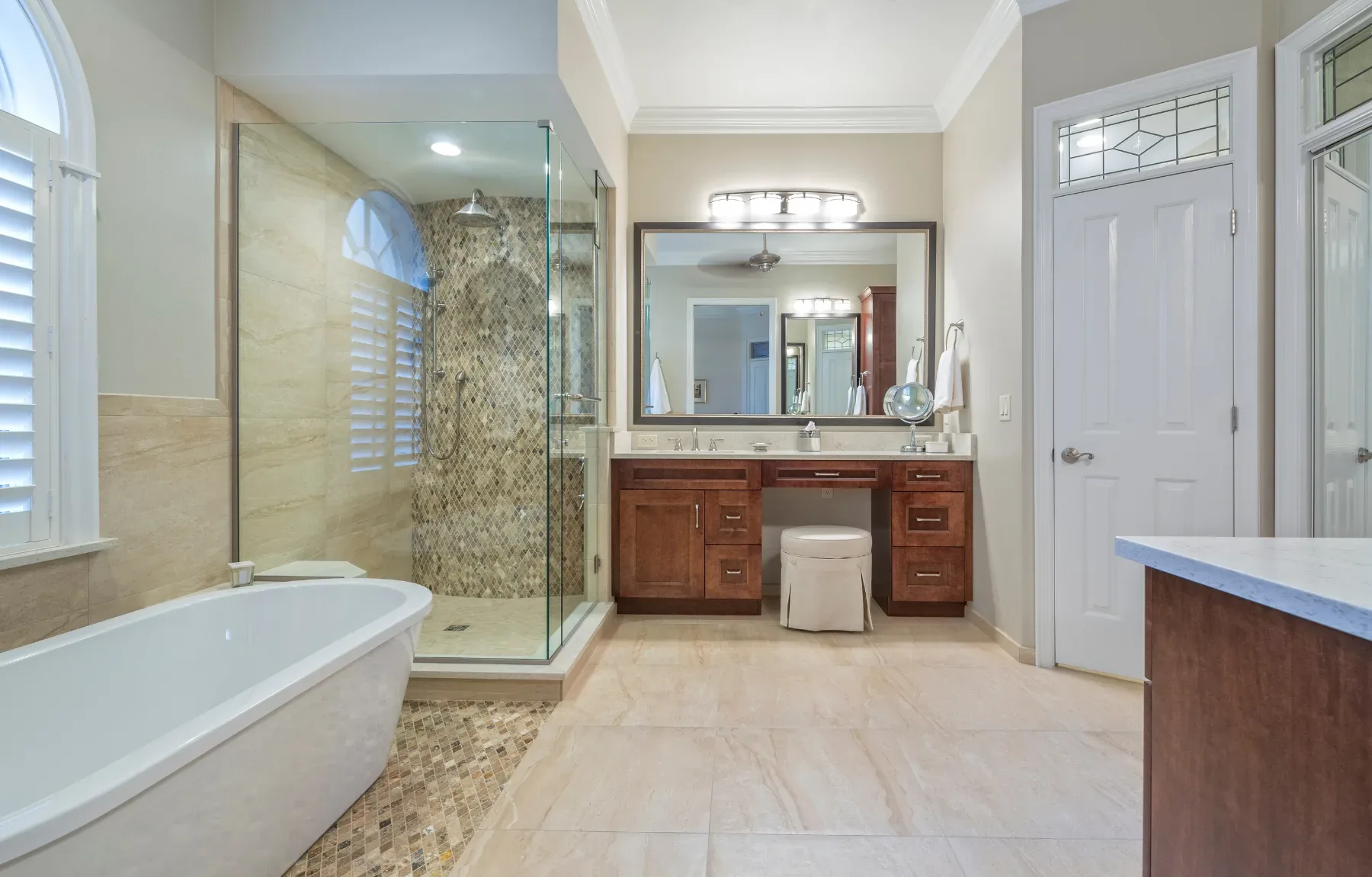 A finished master bathroom with a freestanding white soaking tub at left in front of a large arched window with plantation sh
